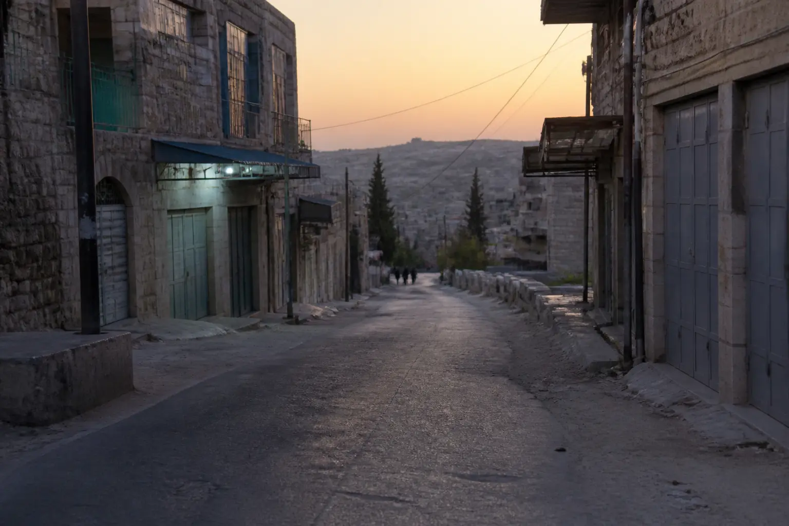 Una tranquila calle de piedra en una ciudad del sur de Cisjordania al atardecer, con tiendas cerradas y un camino polvoriento