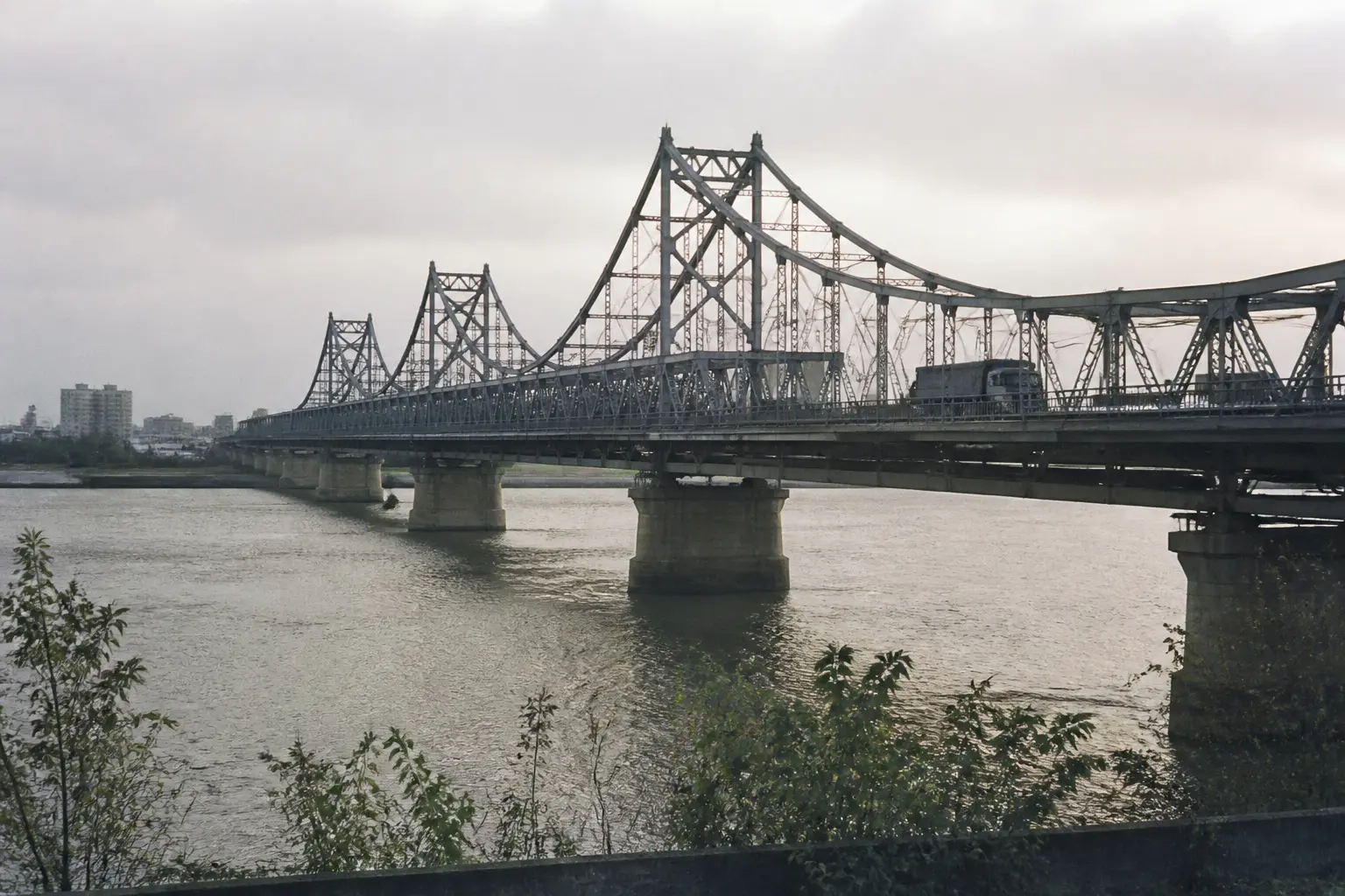 A long steel bridge crossing a wide river between two industrial border cities under pale morning light