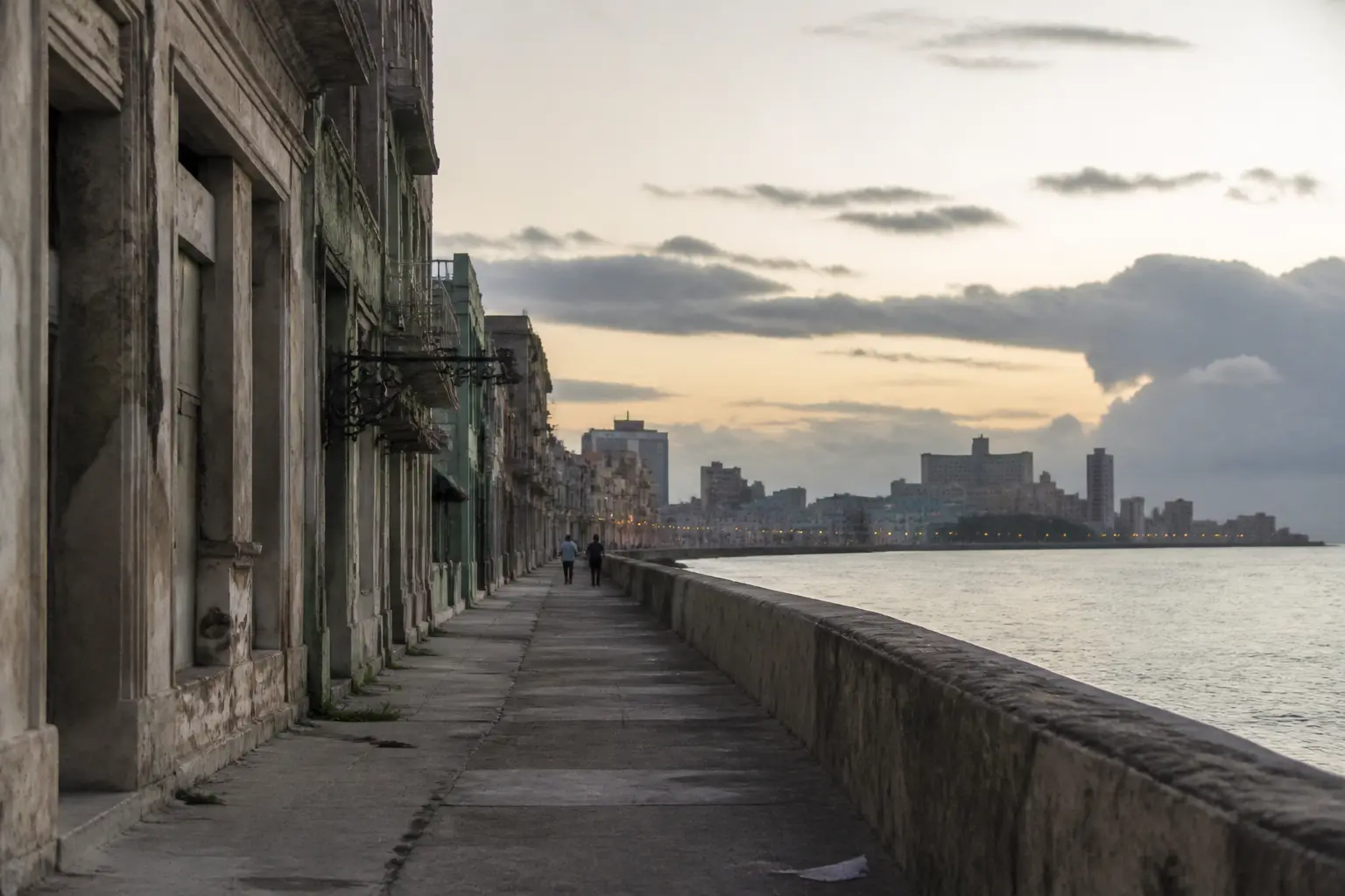 A faded seaside boulevard in a Caribbean city at dusk, with the seawall nearly empty