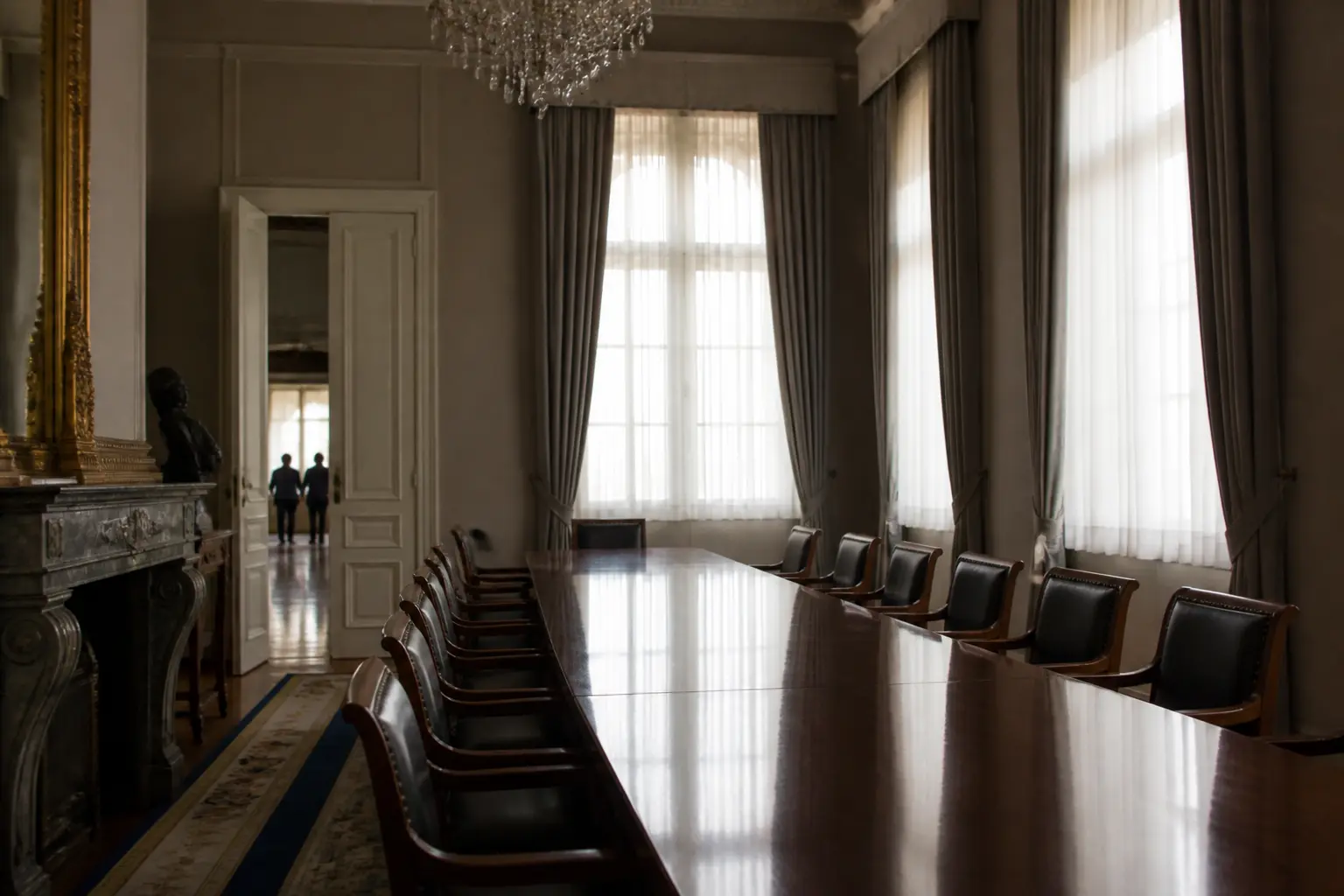 A long empty conference table in a grand room with tall windows and drawn curtains