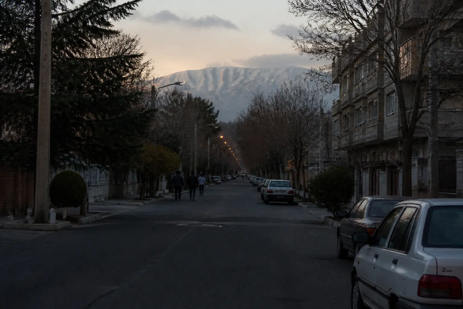 A tree-lined avenue in Tehran at dusk with distant pedestrians and mountains on the horizon